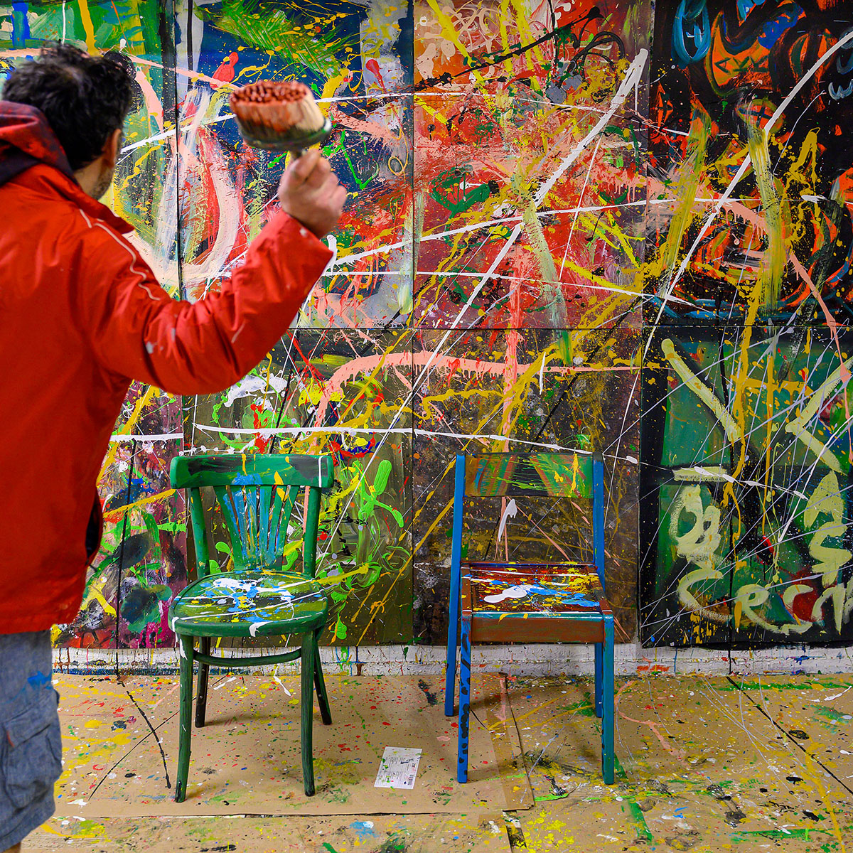 Künstler Eco in roter Jacke wirft Farbe auf zwei mit Farbspritzern bedeckte Stühle vor einer großformatigen, farbintensiven abstrakten Wandmalerei mit Spritzern und Linien