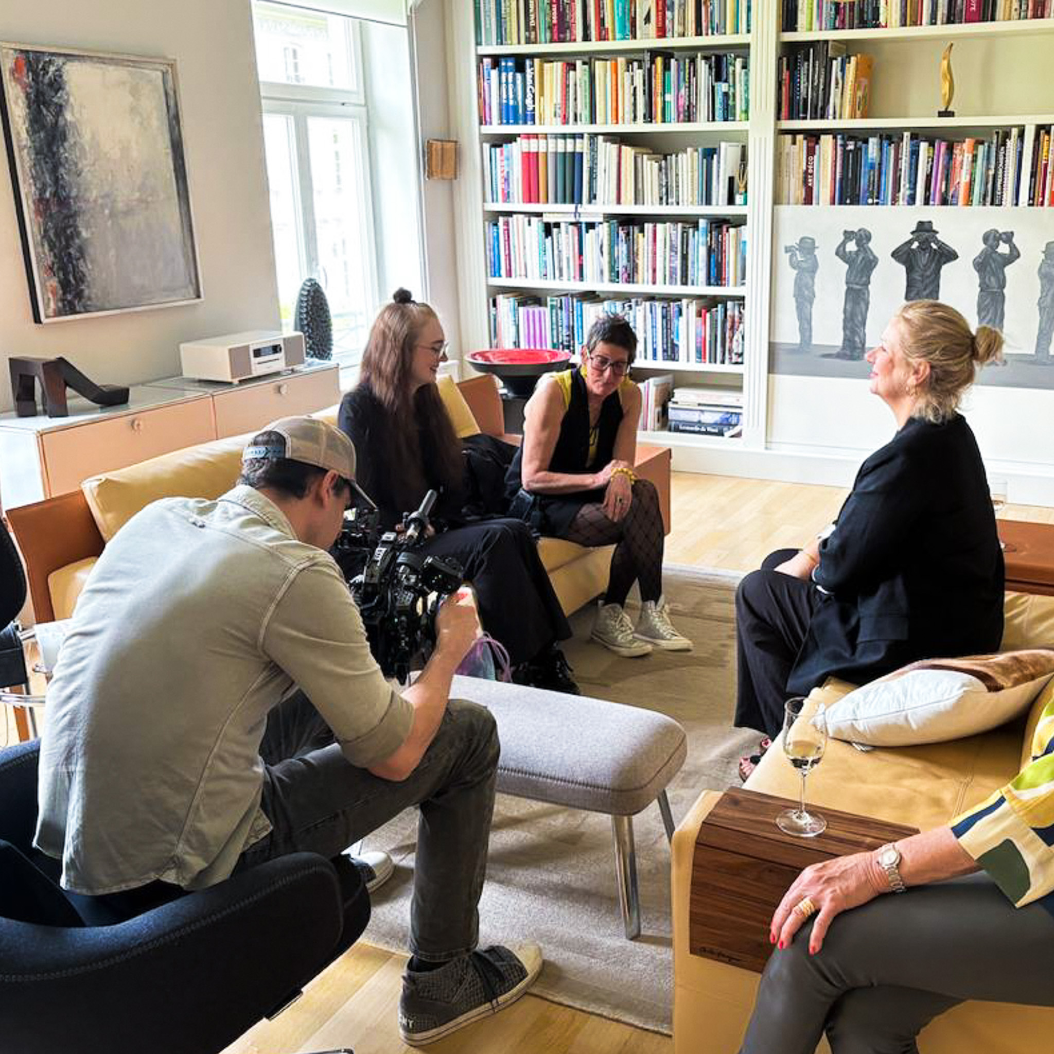 Fünf Personen sitzen in einem Wohnzimmer mit Bücherregal und Kunstwerken und führen ein Gespräch