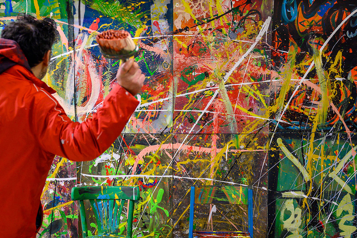 Künstler Eco in roter Jacke wirft Farbe auf zwei mit Farbspritzern bedeckte Stühle vor einer großformatigen, farbintensiven abstrakten Wandmalerei mit Spritzern und Linien