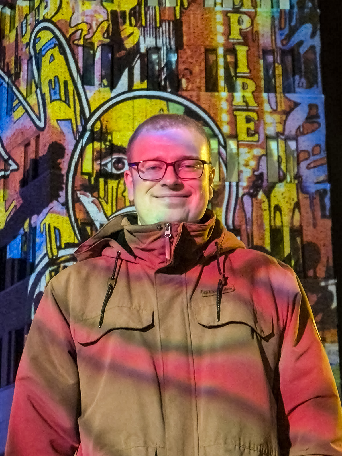 Marius Ohlendorf trägt eine beige Jacke mit Kapuze vor einer bunt beleuchteten Wand mit Graffiti und dem Schriftzug 'EMPIRE'