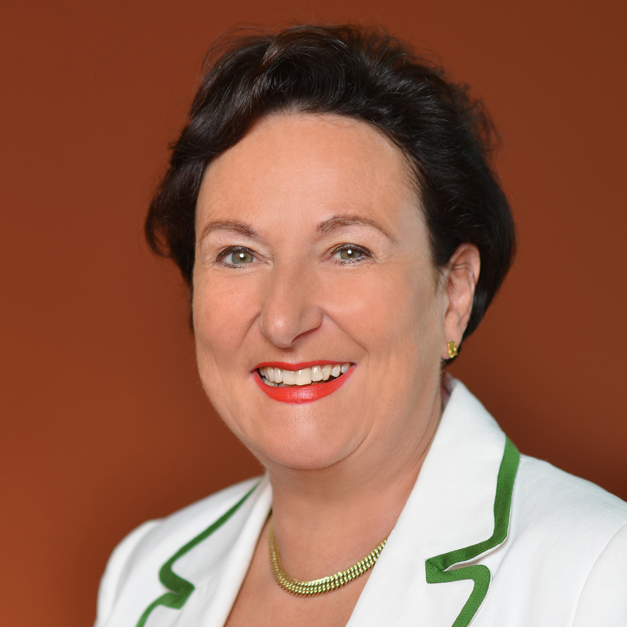 Christine Rother trägt eine weiße Blazerjacke mit grüner Paspelierung, eine grüne Bluse, eine goldene Halskette und goldene Ohrstecker vor orangefarbenem Hintergrund