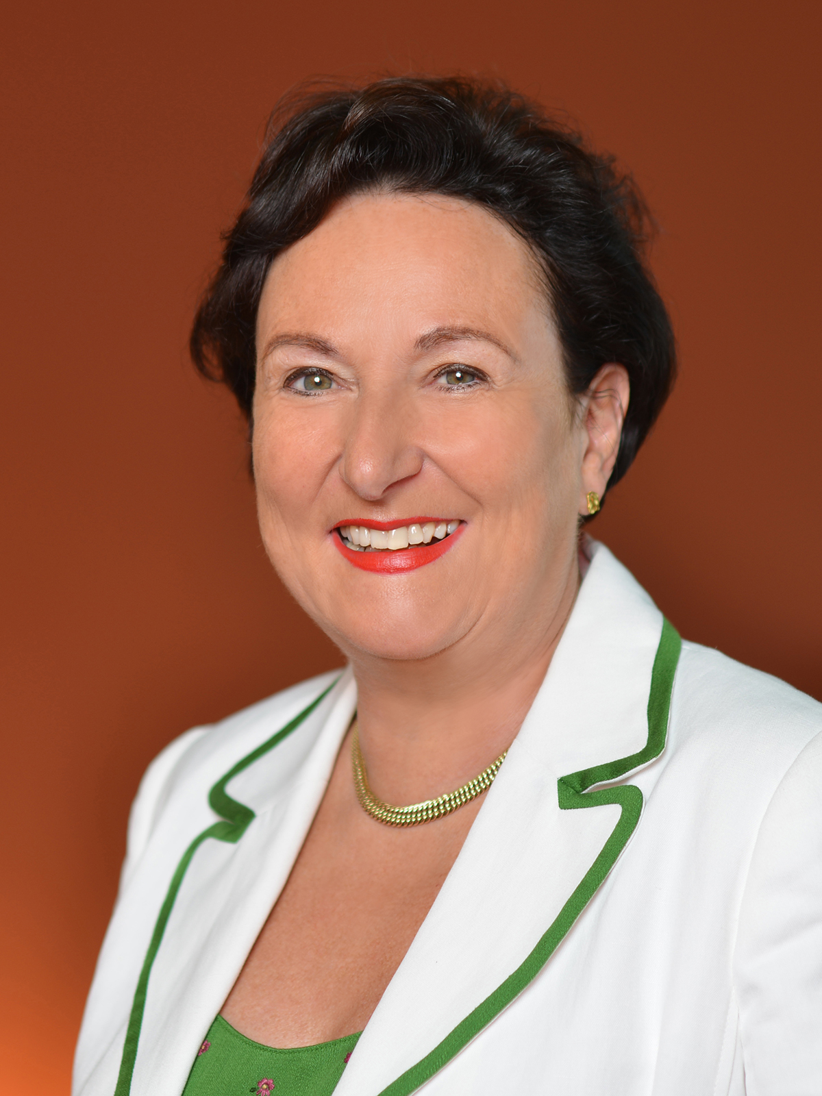 Christine Rother trägt eine weiße Blazerjacke mit grüner Paspelierung, eine grüne Bluse, eine goldene Halskette und goldene Ohrstecker vor orangefarbenem Hintergrund