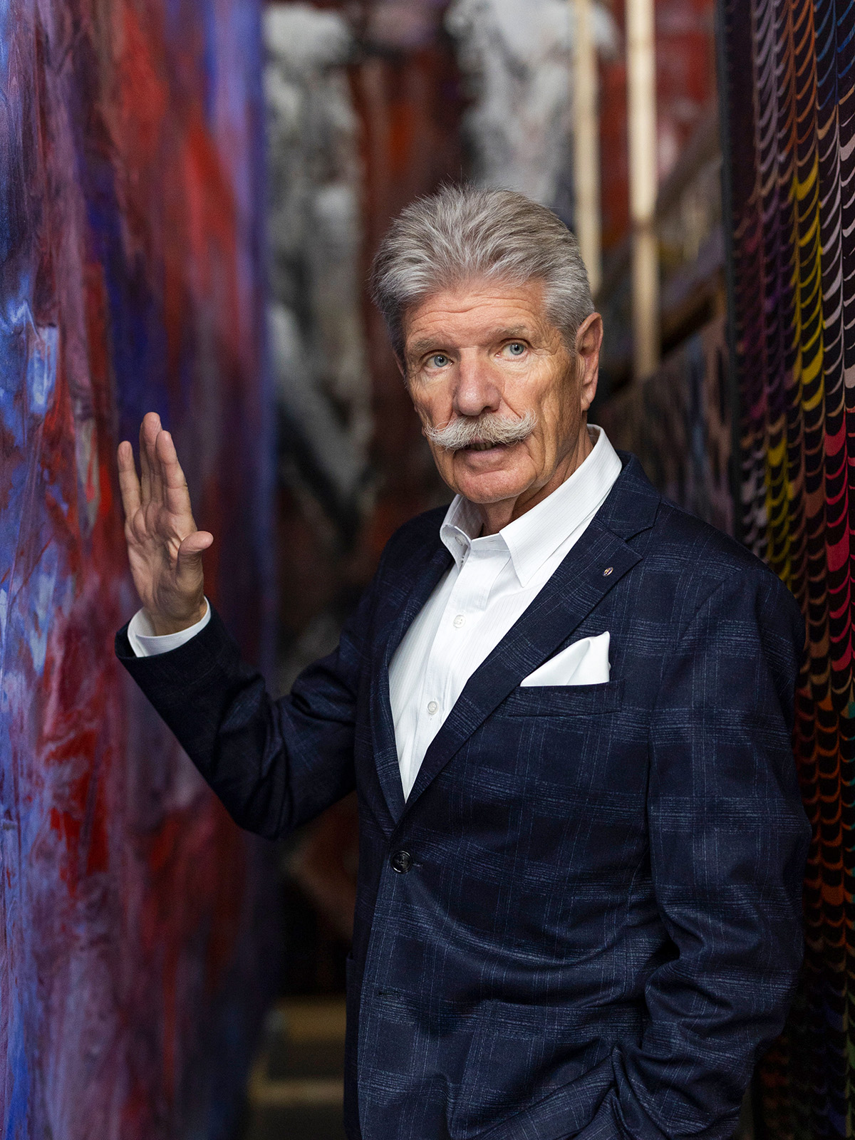 Reinhard Ernst steht in einem schmalen Gang zwischen farbigen Wandgemälden, trägt einen dunklen Anzug mit weißem Hemd und Einstecktuch und hebt die linke Hand leicht an