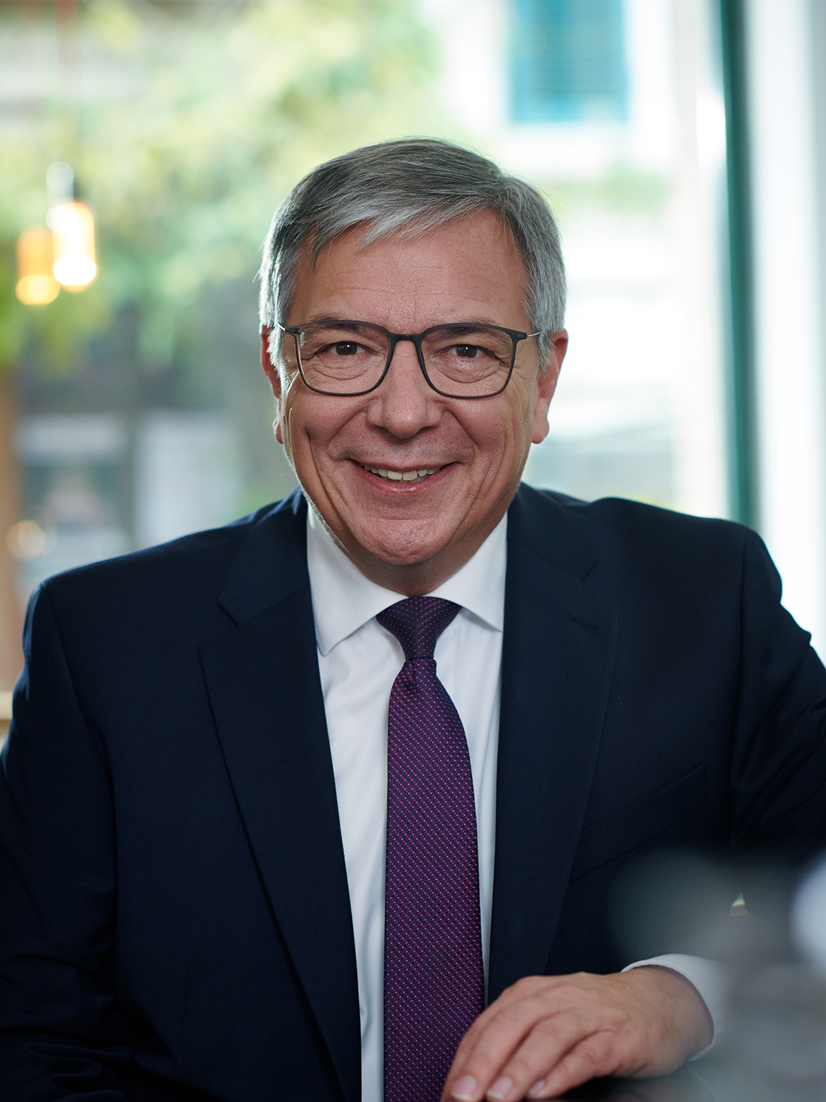 Gert-Uwe Mende sitzt in dunklem Anzug mit weißem Hemd und violetter Krawatte vor einem Fenster mit unscharfem Hintergrund