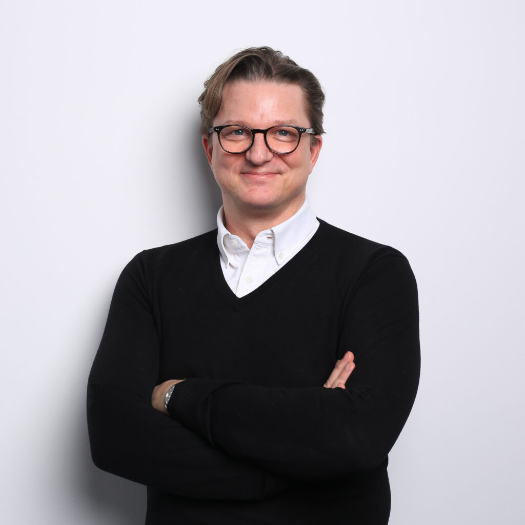 Torsten Anstädt steht mit verschränkten Armen vor weißem Hintergrund und trägt ein weißes Hemd unter einem schwarzen Pullover