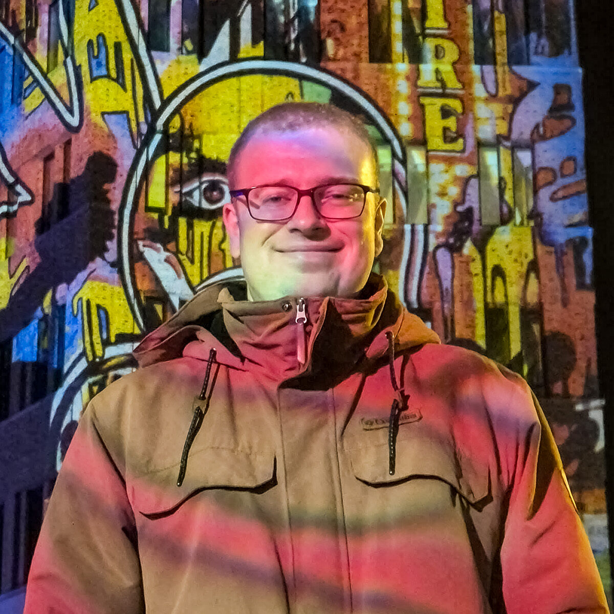 Marius Ohlendorf trägt eine beige Jacke mit Kapuze vor einer bunt beleuchteten Wand mit Graffiti und dem Schriftzug 'EMPIRE'
