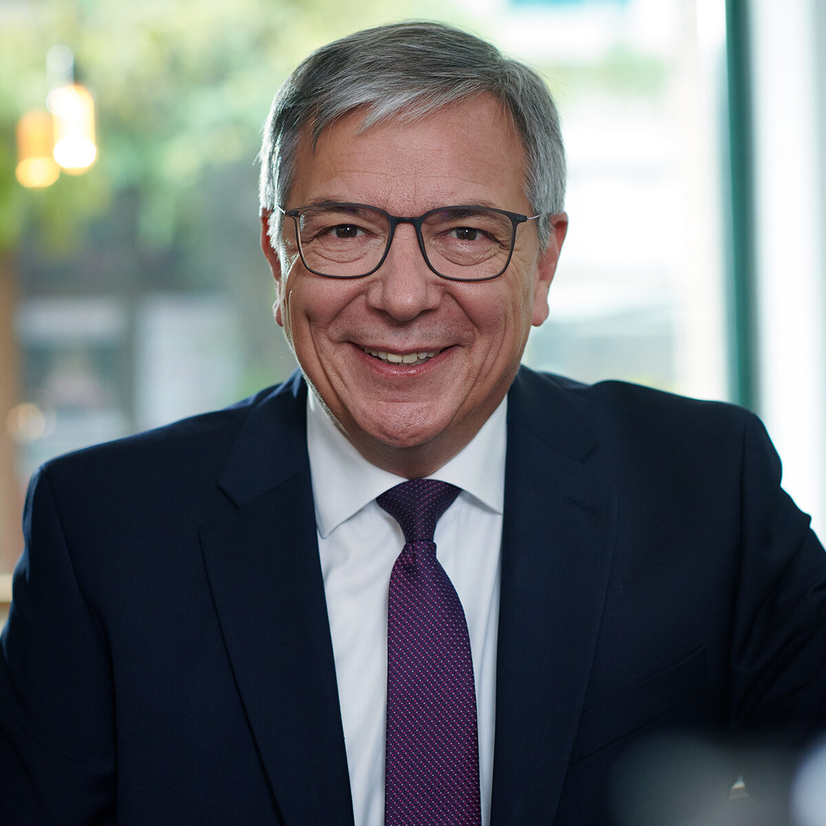 Gert-Uwe Mende sitzt in dunklem Anzug mit weißem Hemd und violetter Krawatte vor einem Fenster mit unscharfem Hintergrund